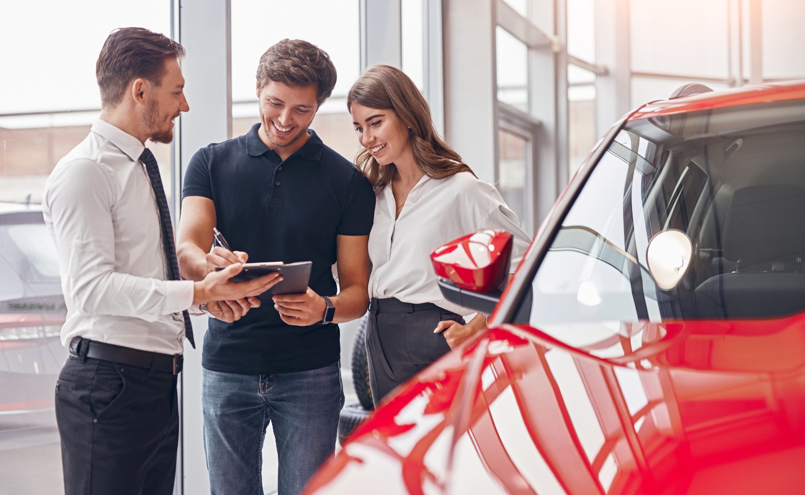 The picture shows three people in a car dealership. On the left, there is a man who appears to be a car salesman. He is wearing a white shirt with a tie and black trousers. He is holding a clipboard and pen, and is showing something on the clipboard to a young couple standing next to him. The couple is standing in the middle of the picture. The man is wearing a black polo shirt and jeans, and the woman is wearing a white blouse and black trousers. They both look happy and are smiling as they look at the clipboard. On the right side of the picture, there is a part of a red car visible. It seems to be a new car displayed in the dealership. The car's front door is open. The background shows large windows, and it seems to be a bright day outside.